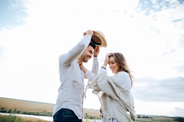 Happy couple in love hugging, kissing and smiling against the sky in field. Hat in girl's hand