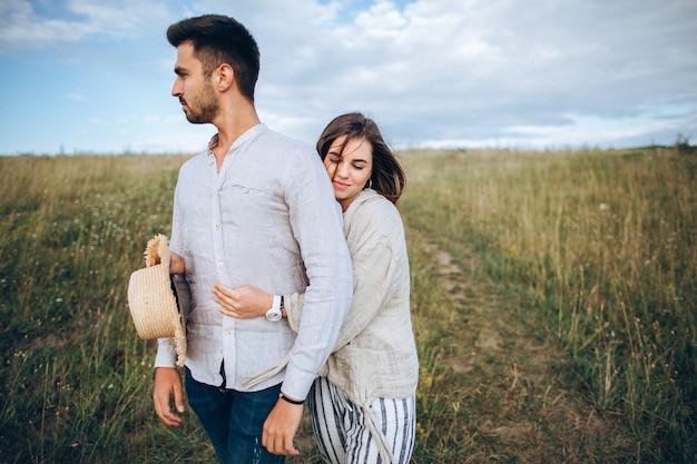 Happy couple in love hugging, kissing and smiling against the sky in field. Hat in girl's hand