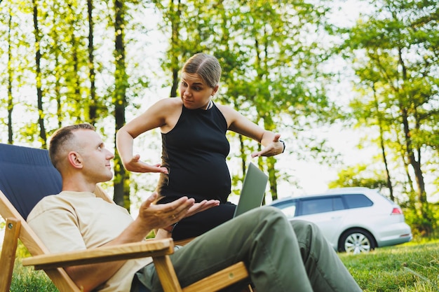 A happy couple relaxes in folding chairs near the car a man uses a laptop for work or checking social networks his wife treats him to a croissant Pregnant woman with man in nature