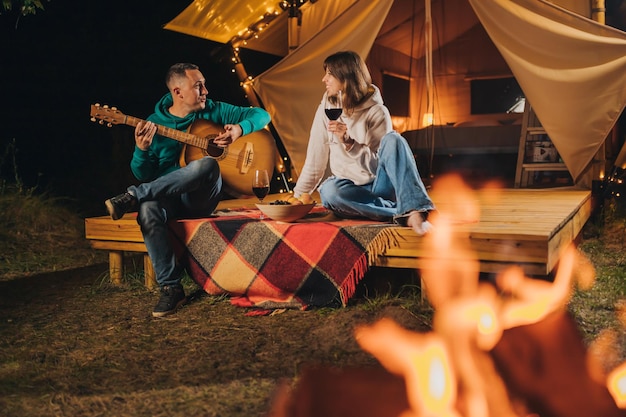 Happy couple relaxing in glamping on autumn evening drinking wine and playing guitar near cozy bonfire Luxury camping tent for outdoor recreation and recreation Lifestyle concept