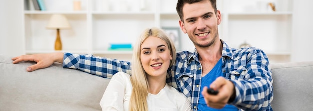 The happy couple sit on the sofa and hold the remote control