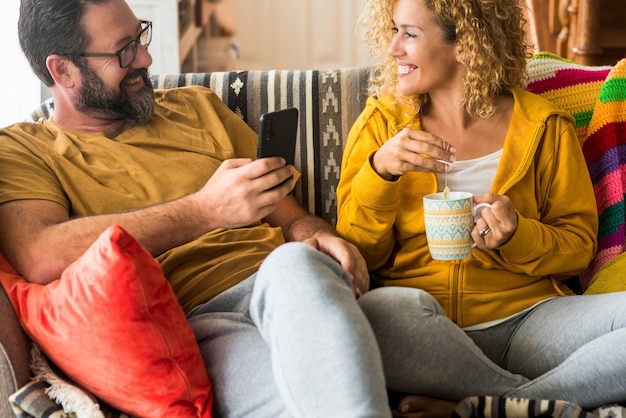 Happy couple sitting on sofa and looking at each other while drinking coffee and using mobile phone at home. Loving couple spending leisure time together at home. Couple looking face to face.