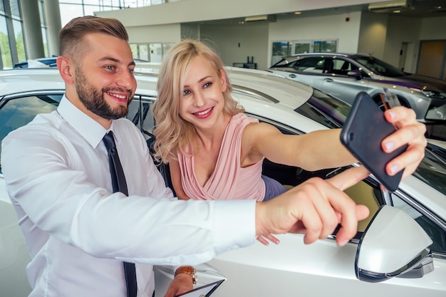 Happy couple stand in front of new car man with his wife eyes making selfie on smartphone camera in showroomsurprised gift automobile