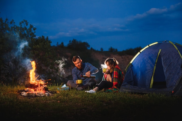 Happy couple travellers sitting together beside campfire and glowing tourist tent