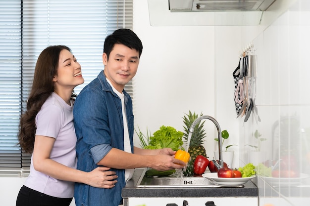 Happy couple washing sweet pepper and vegetables in the sink in the kitchen at home woman is hugging man