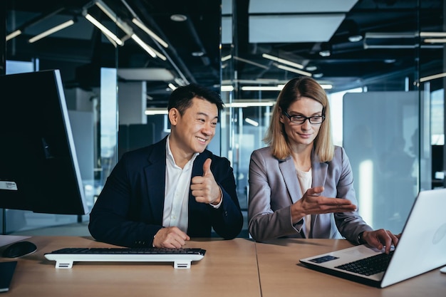 Happy coworkers discuss project strategy by looking and pointing at laptop pc computer monitor screen multiethnic business team in the office confident mature asian man explaining young female