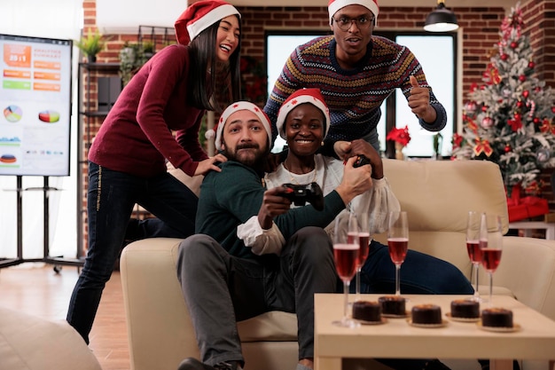 Happy coworkers having fun with video games at event in festive office decorated with xmas tree and ornaments. Playing competition on console and celebrating christmas holiday festivity.