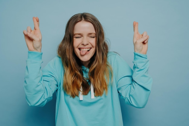 Happy crazy teenage girl with wavy ombre hair eyes closed fingers crossed sticking tongue out standing against blue wall in studio carefree and having fun