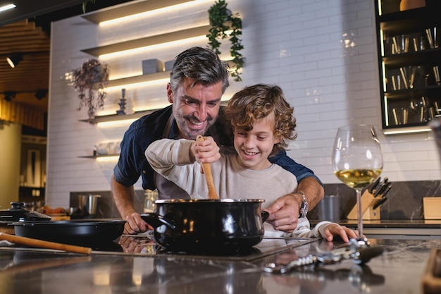 Happy curly haired boy stirring hot dish in saucepan and helping middle aged father with cooking while spending time together in kitchen at home