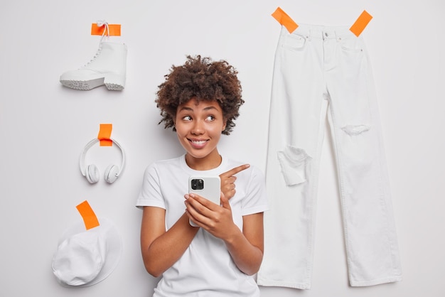 Happy curly haired young woman dressed in casual t shirt points index finger demonstrates different clothes uses mobile phone for online shopping isolated over white background Check this out