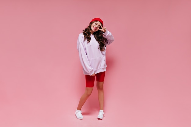 Happy curly woman in red hat shows peace sign