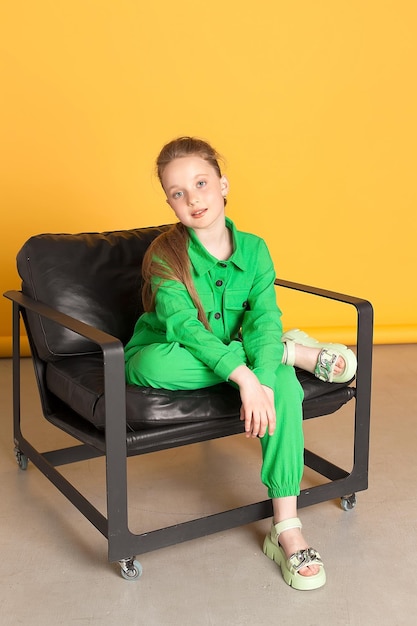 A happy curlyhaired girl with long hair in a light green jumpsuit She is sitting on a black armchair against the yellow background of the studio Childhood fashion Full length copy space