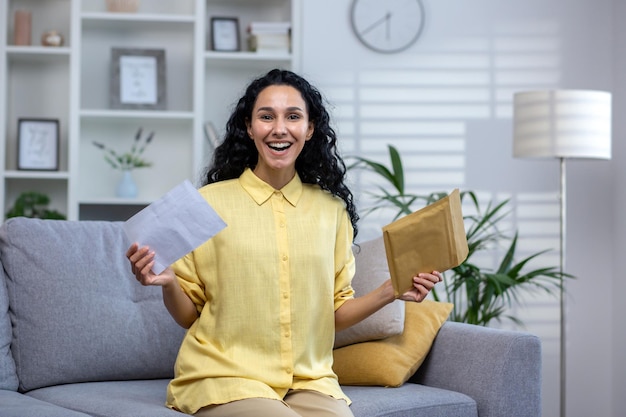 Happy curlyhaired hispanic woman sitting at home on sofa in living room holding open envelope cute