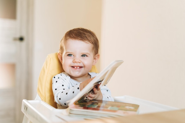 Happy cute baby girl in booster seat reading children book at the home