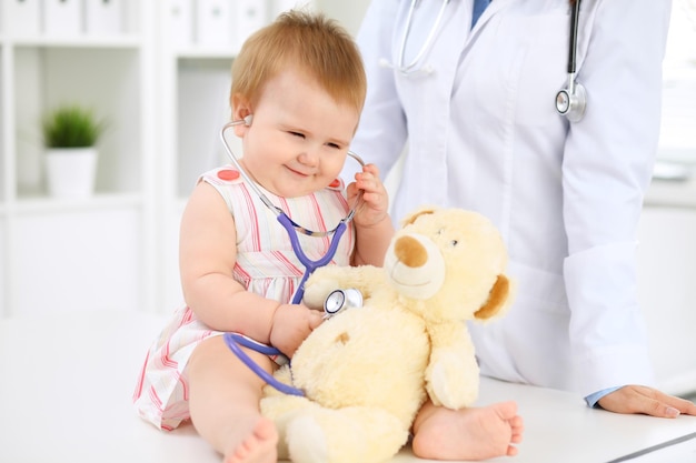 Happy cute baby at health exam at doctor's office Toddler girl is sitting and keeping stethoscope and teddy bear