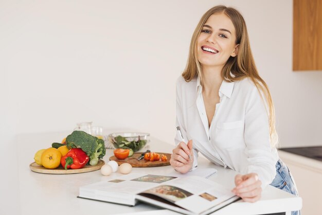 A happy cute blonde woman is writing something down and looking at a recipe book in the kitchen