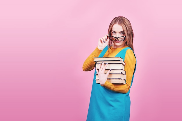 Happy cute girl in glasses holding in hands a pile of books isolated on colorful pink 