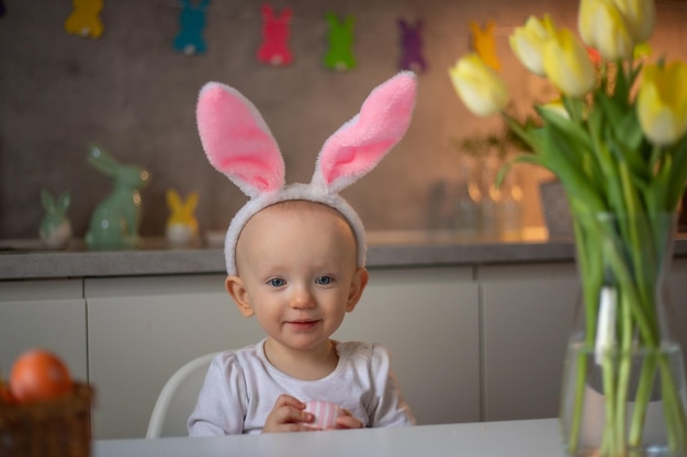 Happy cute little baby girl wearing bunny ears on easter day sitting at the table in the kitchen