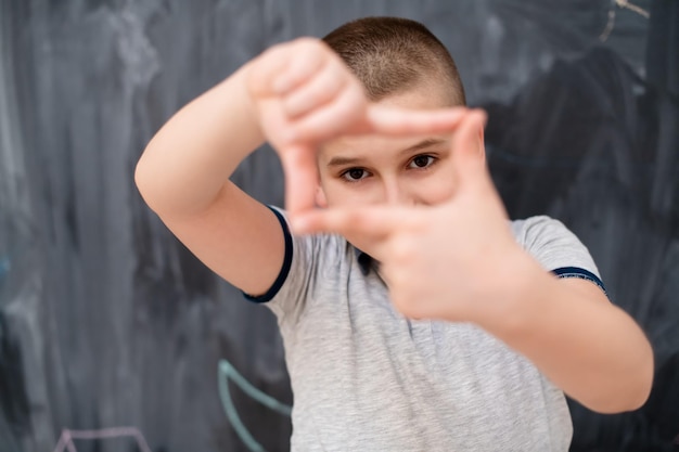 happy cute little boy having fun making hand frame gesture while standing in front of black chalkboard