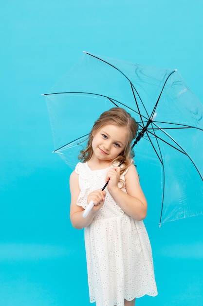 Happy cute little girl in a cotton white dress holding a transparent umbrella on a blue background in the studio and smiling and fooling around space for text