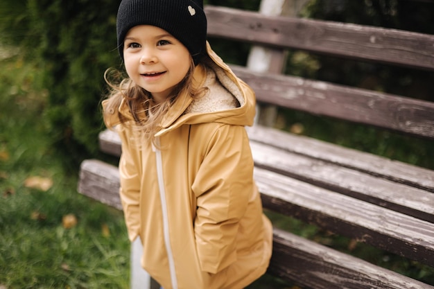 Happy cute little girl in jacket and hat posing to photographer outdoor by the bench little model