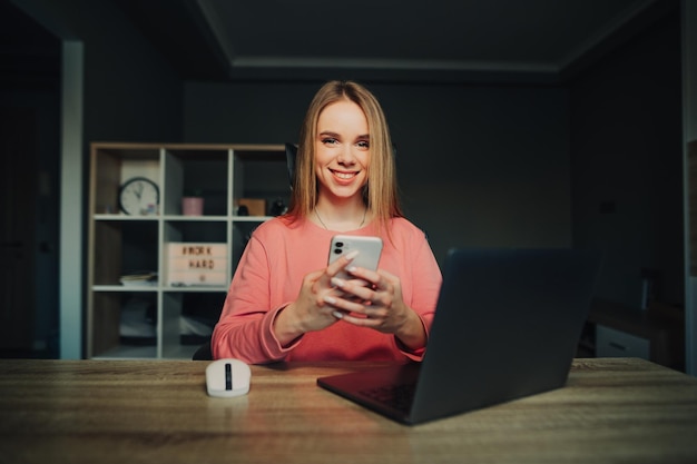 Happy cute woman sitting at home at work with a laptop and using a smartphone