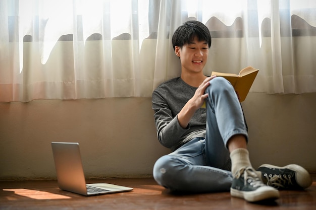 Happy and cute young Asian man sits on the floor in his room and enjoys reading a book