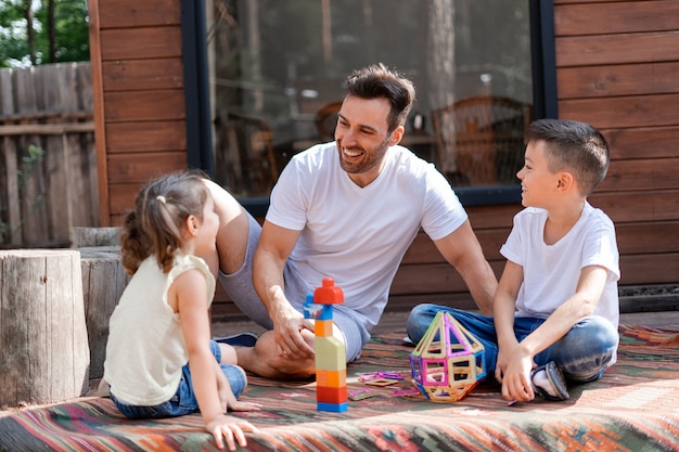 Photo happy dad or babysitter has fun with little girl and boy, plays with them in the yard sitting on the carpet near the wooden house and helps them gather children's toys