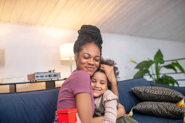 happy darkskinned young mother with little cute daughter with gift hugging with closed eyes