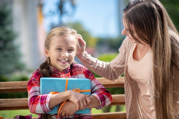 Happy daughter hoding present box, sitting with her mom on park bench