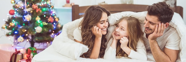 The happy daughter and parents lay on the bed near the christmas tree