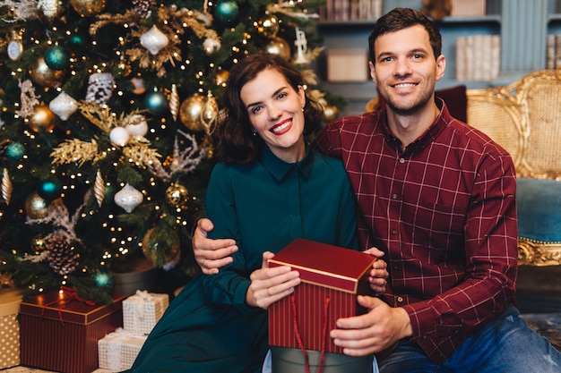 Happy delightful couple in love, embrace each other, sit near decorated New Year tree, hold present