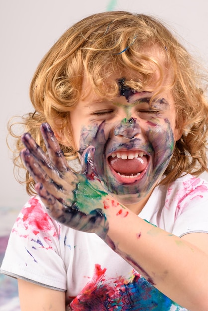 Happy dirty boy covered with colorful paints having fun while sitting in light studio with closed eyes on white background