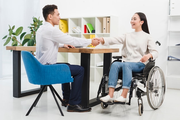 Happy disabled young woman sitting on wheel chair shaking hands with male colleague in the office