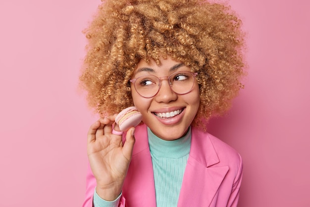 Happy dreamy young woman with curly bushy hair looks away has sweet tooth holds tasty macarroon eats unhealthy food dressed formally isolated over pink background Female eats yummy dessert