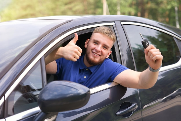 Happy driver young man driving his car show keys smiling show thumb up like  from automobile window