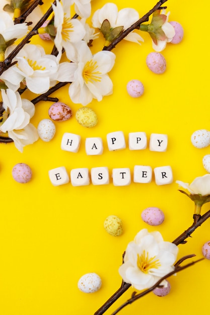 Happy Easter. Cubes with text on yellow background. Spring brunches with white flowers and candy, chocolate easter eggs. Minimalistic flat lay.