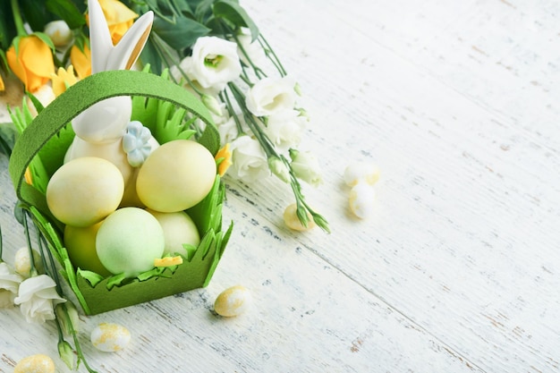 Happy Easter Easter eggs and rabbit in green basket on white old wooden table with white and yellow roses Spring Happy Easter holiday card Easter background with copy space Top view