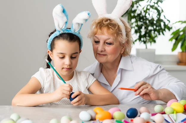 Happy easter. A grandmother and her granddaughter painting Easter eggs. Happy family preparing for Easter. Cute little child girl wearing bunny ears on Easter day.