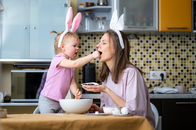 Happy easter Joyful family mom and daughter wearing bunny ears headbands gathering at table in kitchen and preparing baking cake Easter together