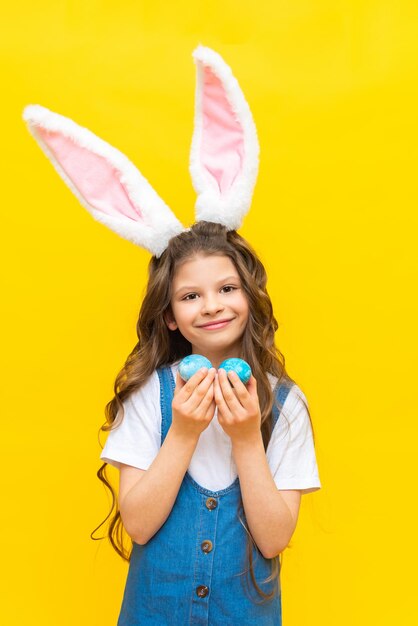 Happy Easter A little girl holds two painted eggs for the holiday A charming baby with rabbit ears on a yellow isolated background