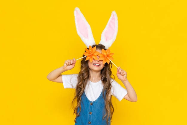 Happy Easter A little girl is happily celebrating a spring holiday A child with ears on his head depicts a rabbit A little lady holds origami flowers on a yellow isolated background