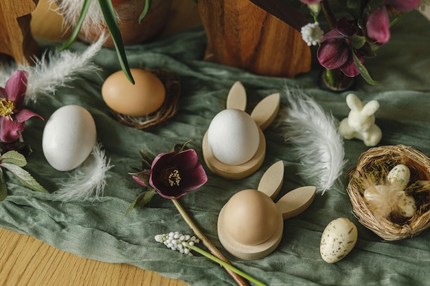 Happy Easter Stylish wooden bunny ears and natural eggs spring flowers feathers and nest on rustic table in room Easter still life Festive arrangement and decor in farmhouse top view