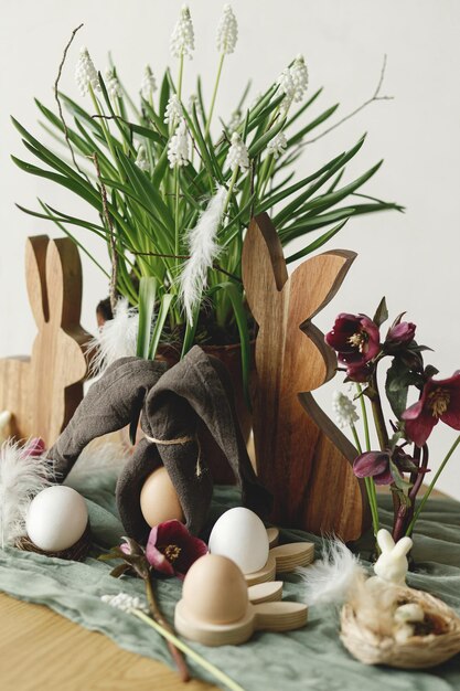 Happy Easter Stylish wooden bunny spring flowers natural eggs and napkin bunny ears on rustic table in room Easter still life Festive arrangement and decor in farmhouse