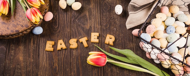 Happy Easter surface, Lettering from cookies, tulips and colorful quail eggs in metal basket on wooden surface. Banner