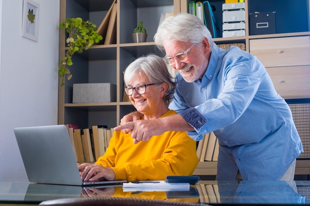 Happy elderly 60s couple sit rest on couch at home pay household expenses online on computer smiling mature 50s husband and wife clients hold documents make payment on internet banking servicexA