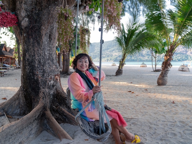 happy elderly asian woman in colorful dress sit and relax on swing under big tree on seaside beach