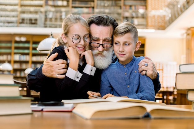 Happy elderly bearded man, grandfather and his cute grandson and granddaughter are sitting at the table in vintage old library and reading books