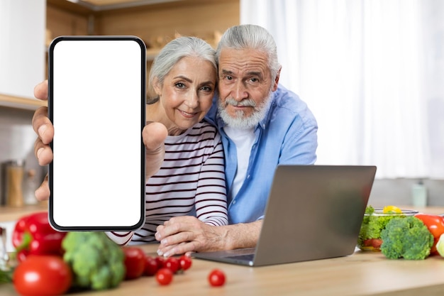 Happy Elderly Couple Showing Big Blank Mobile Phone While Posing In Kitchen