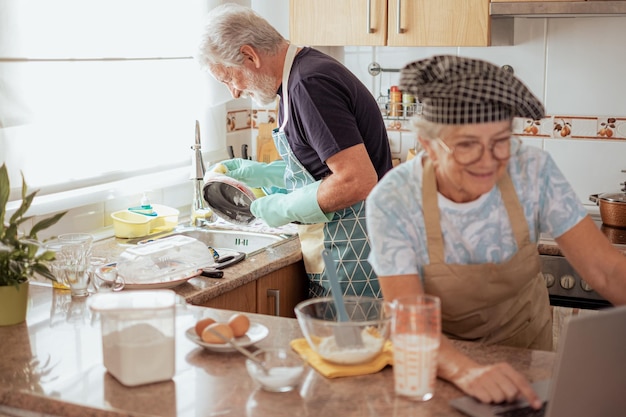 Happy elderly couple working together in the home kitchen husband washing dishes wife preparing a homemade cake looking at web pages by laptop for the right recipe